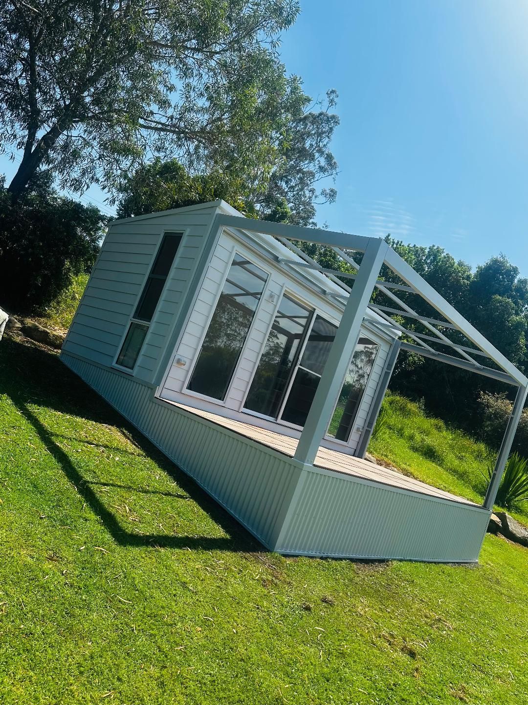 A light-blue cabin with large windows and a covered deck sits on a grassy hill, under a bright blue sky.