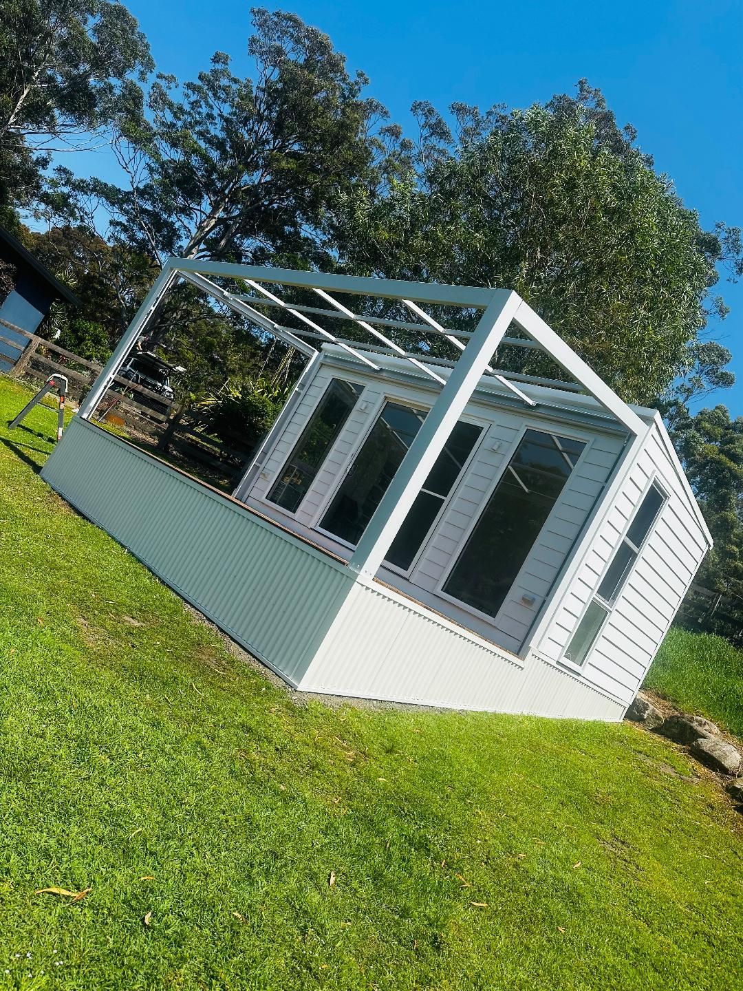 White greenhouse with glass doors and windows, set on a grassy hill, surrounded by trees.