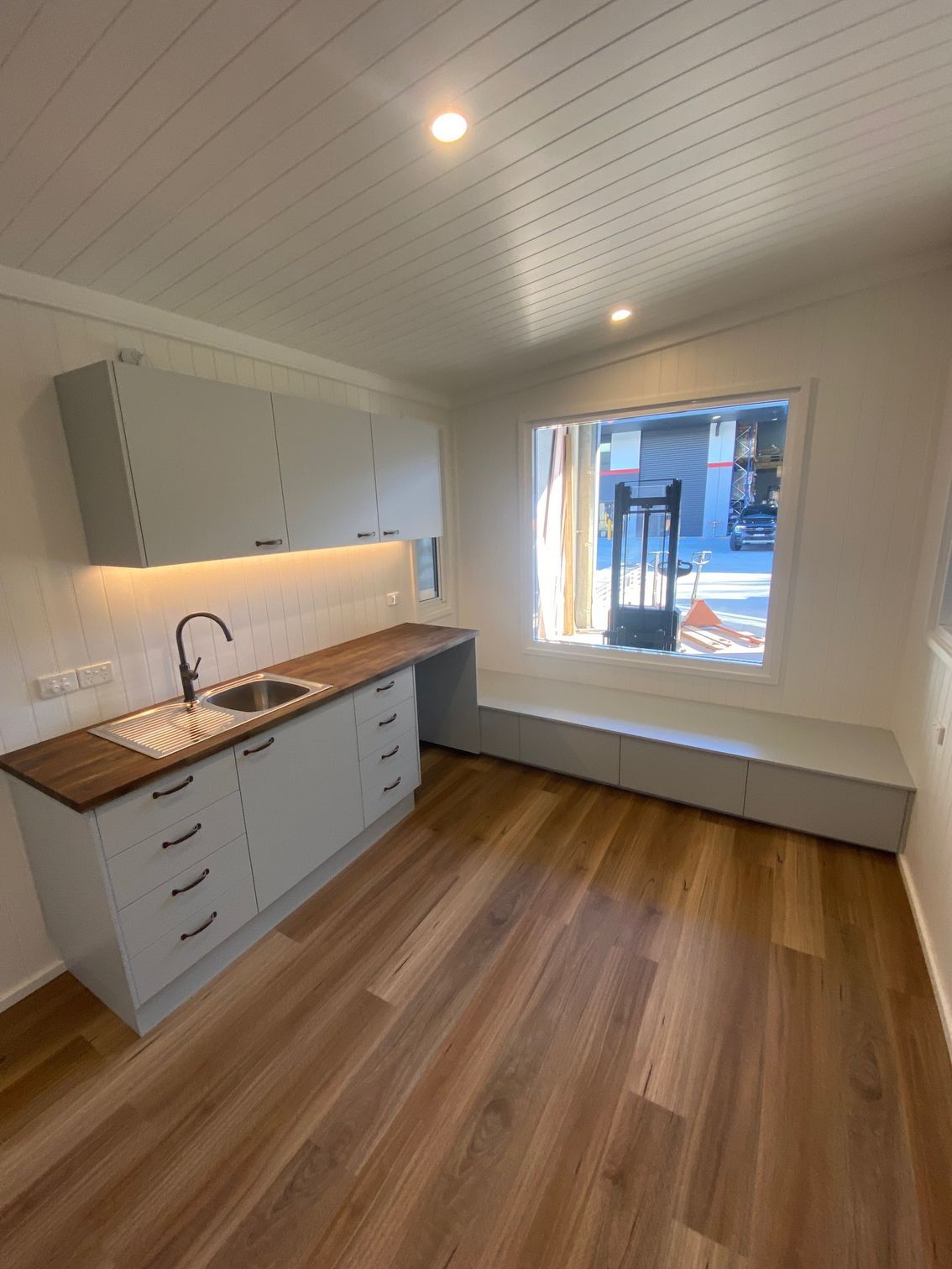 Modern kitchen with light gray cabinets, wood-look flooring, and a window seat in a bright, minimalist room.
