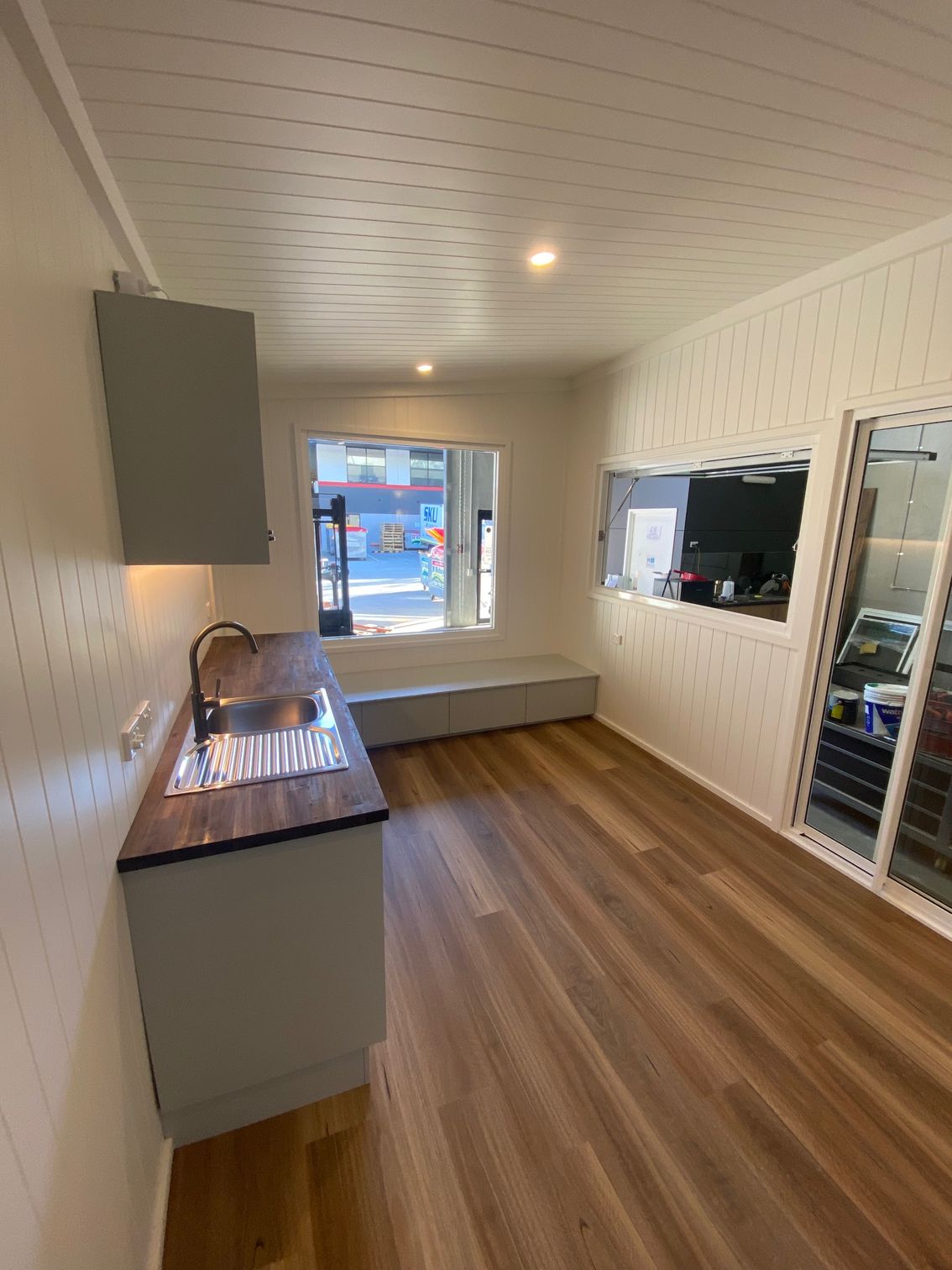 A small, modern kitchen featuring grey cabinets, a wooden countertop with a sink, and light wood flooring in a bright room.