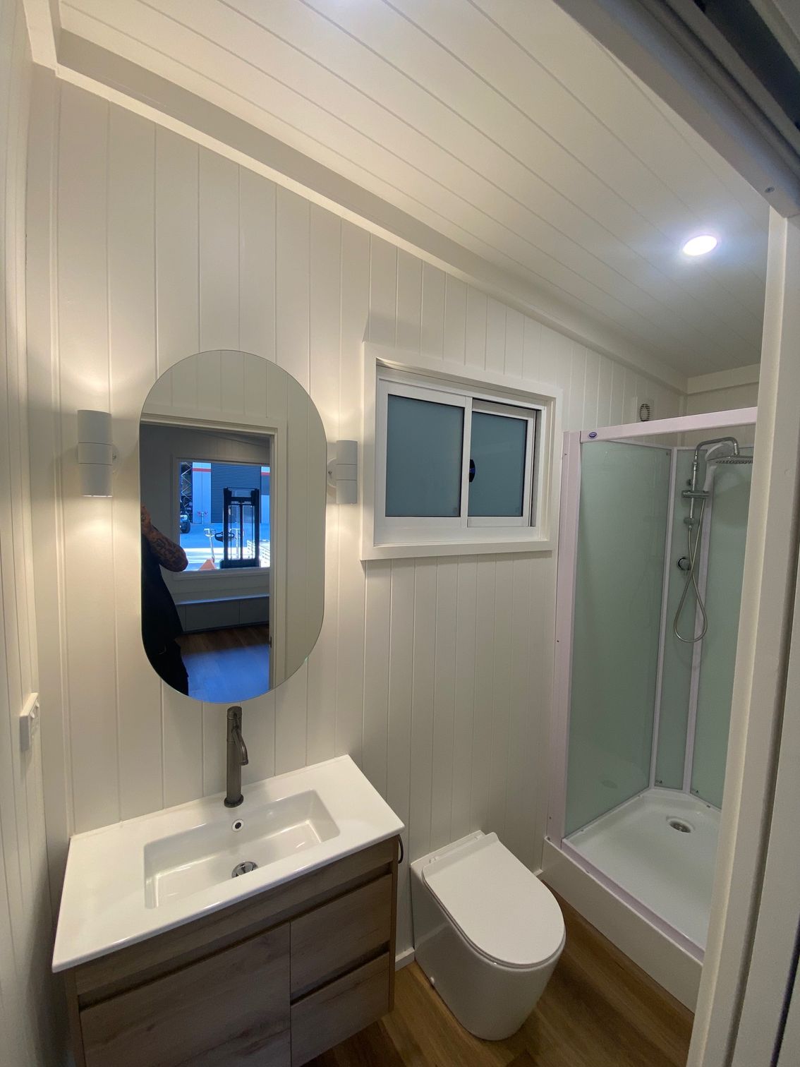 A modern, white-paneled bathroom featuring a wood vanity with a vessel sink, oval mirror, wall-mounted toilet, and shower.