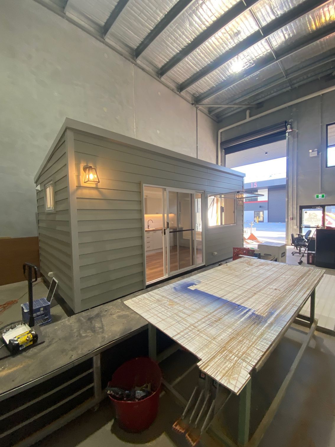 A gray prefab tiny house sits inside a warehouse workshop next to a large work table.