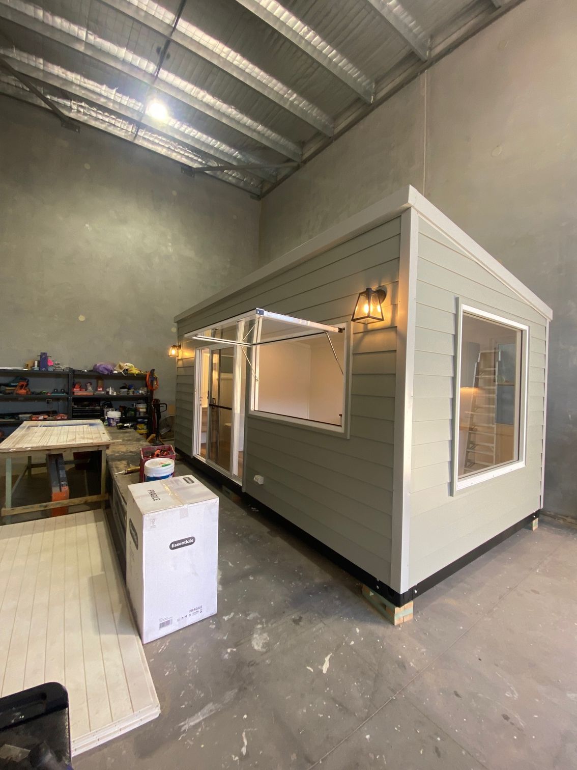A light green shed with a serving window and exterior lighting sits on blocks inside a warehouse.