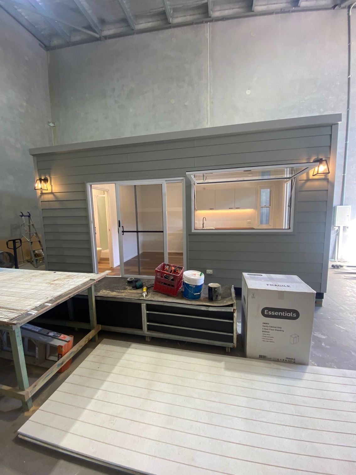 A grey, modular outdoor cabin under construction in a warehouse with a sliding glass door and a rectangular window.