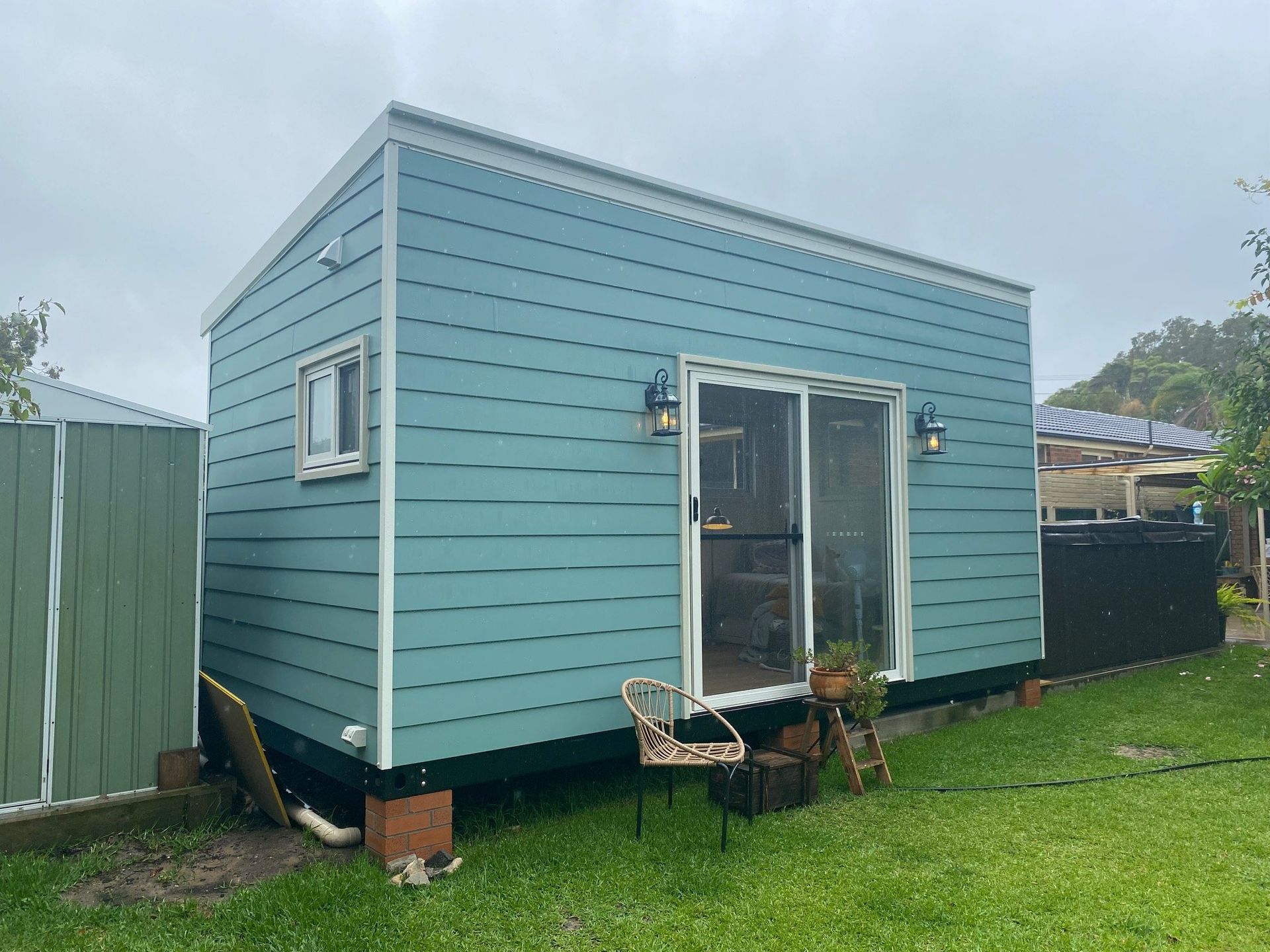 Blue tiny house with a sliding glass door and small window, sitting on cinder blocks in a yard.
