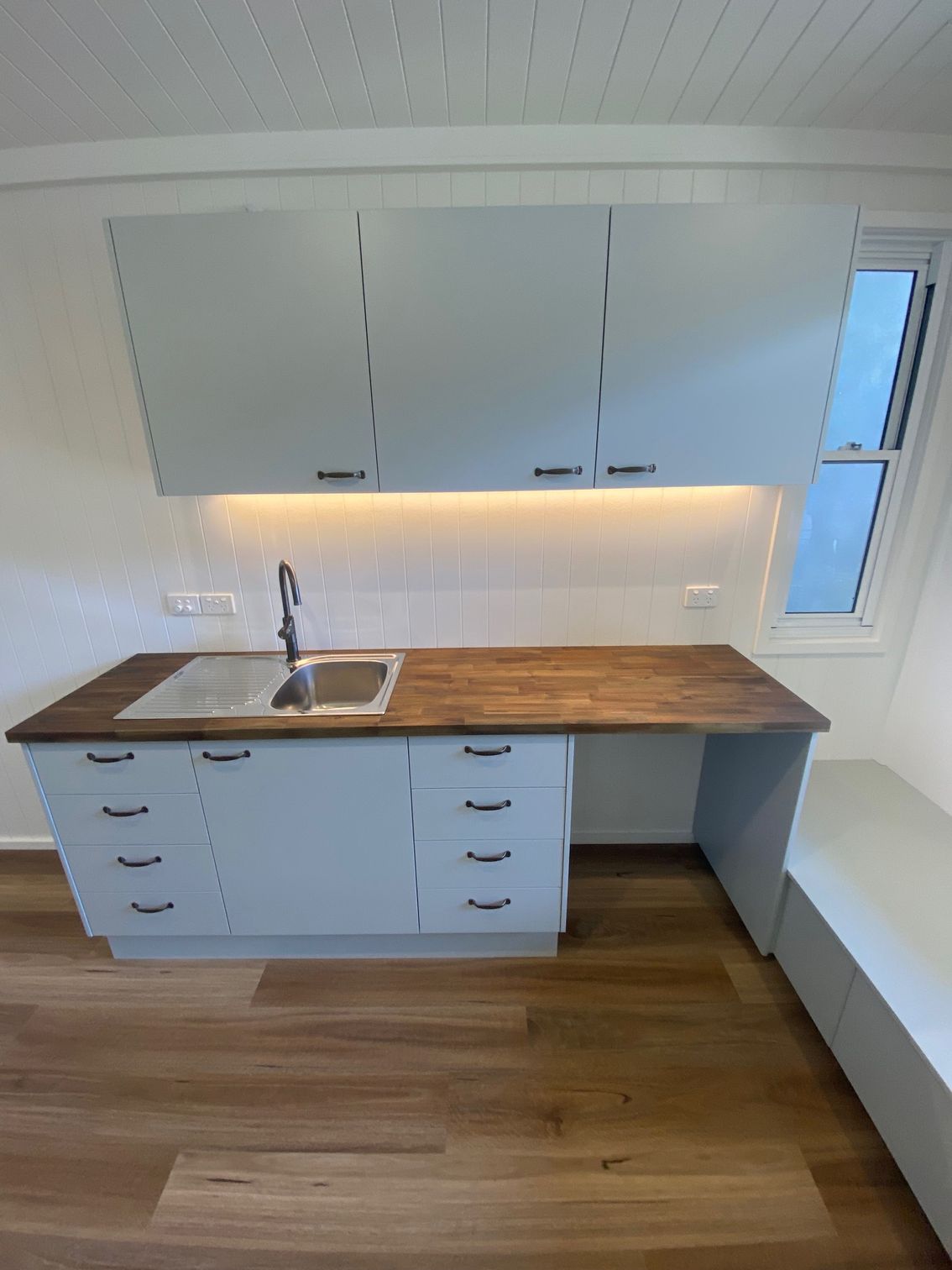 A kitchen area with a wood countertop, grey cabinets, a stainless steel sink, and under-cabinet lighting.