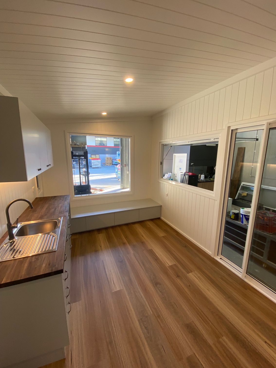A modern, bright indoor room with wood-look flooring, a small kitchenette, built-in bench seating, and glass sliding doors.