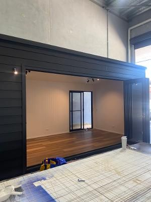 A modern, dark-clad structure with wide open folding doors revealing an interior with wood flooring and a glass door.