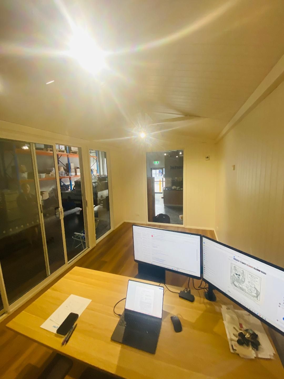 Office with two monitors, a laptop, and a desk. A window and door are visible. Bright overhead light.