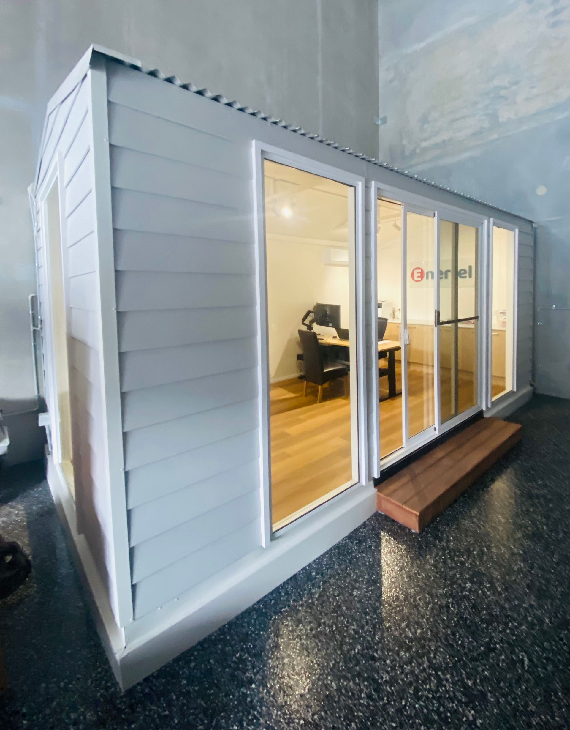 Gray shed with glass doors, wood trim, and a small step. Inside, an office setup with a desk is visible.