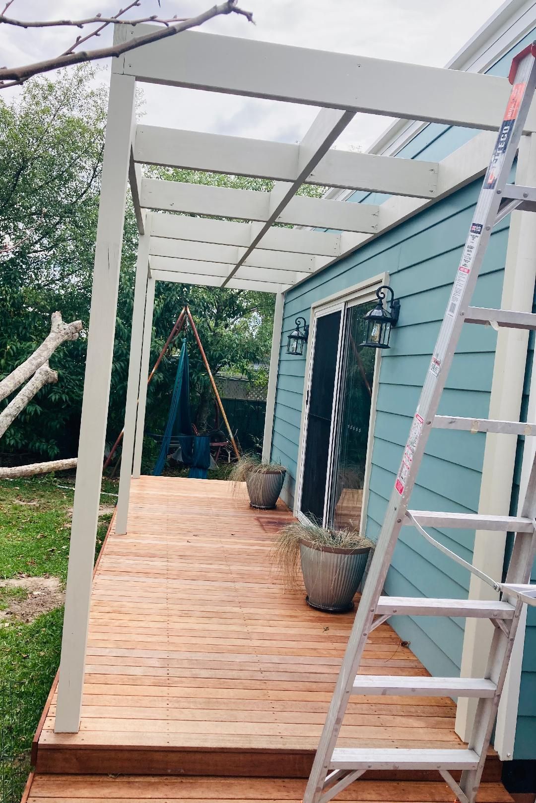 Wooden deck and pergola attached to a blue house with a ladder.