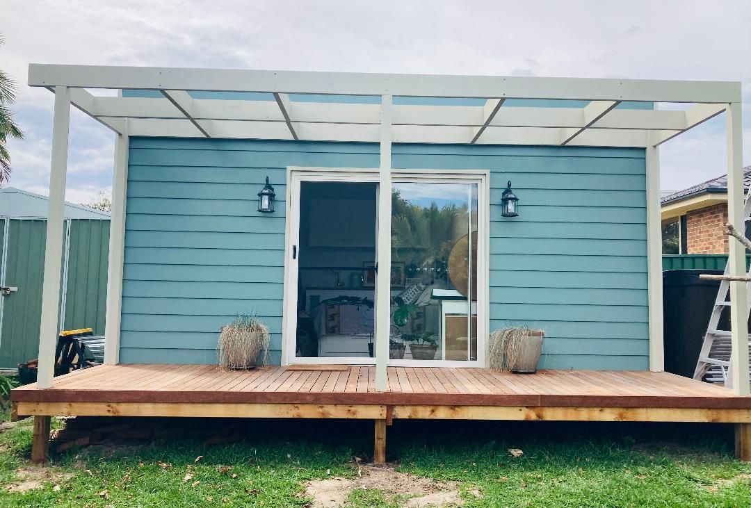Blue shed with white pergola, wooden deck, glass sliding doors, and two sconces.
