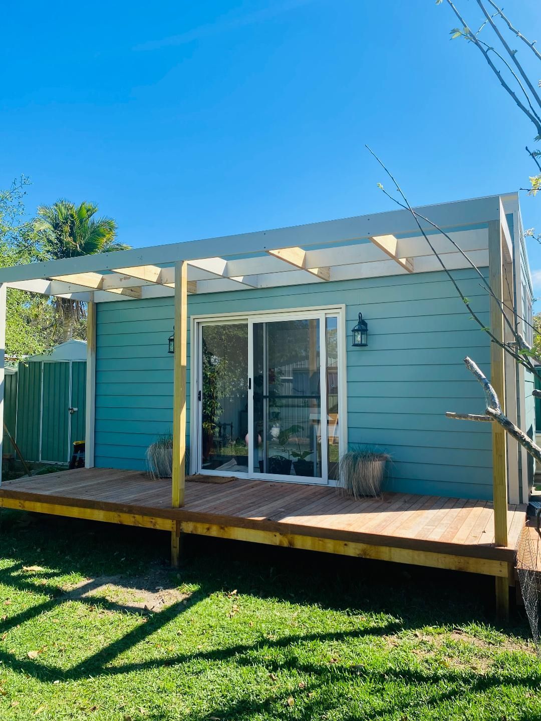 Teal backyard shed with wooden deck, pergola, and sliding glass door on a sunny day.