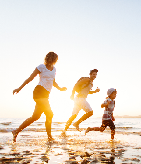 Family running on a beach at sunset; woman, man, and child splashing in the water.