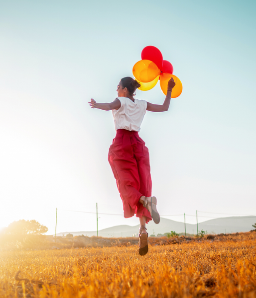 Woman jumps in a field holding red and yellow balloons; arms outstretched, joyful.