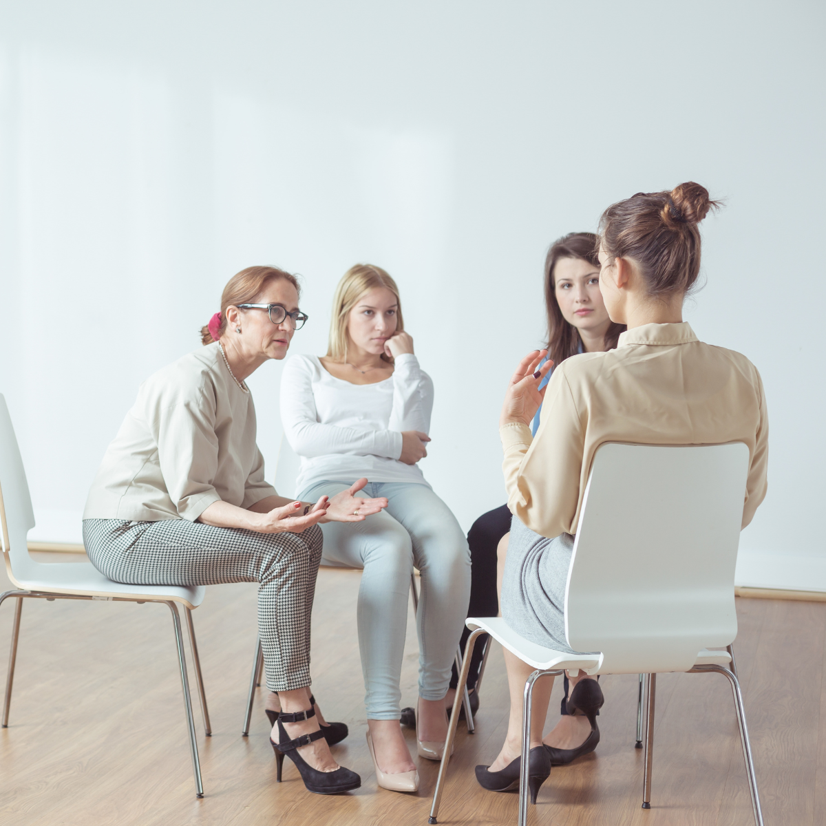 Four women in a circle, one talking, in a bright room. One woman gestures, others listen thoughtfully.