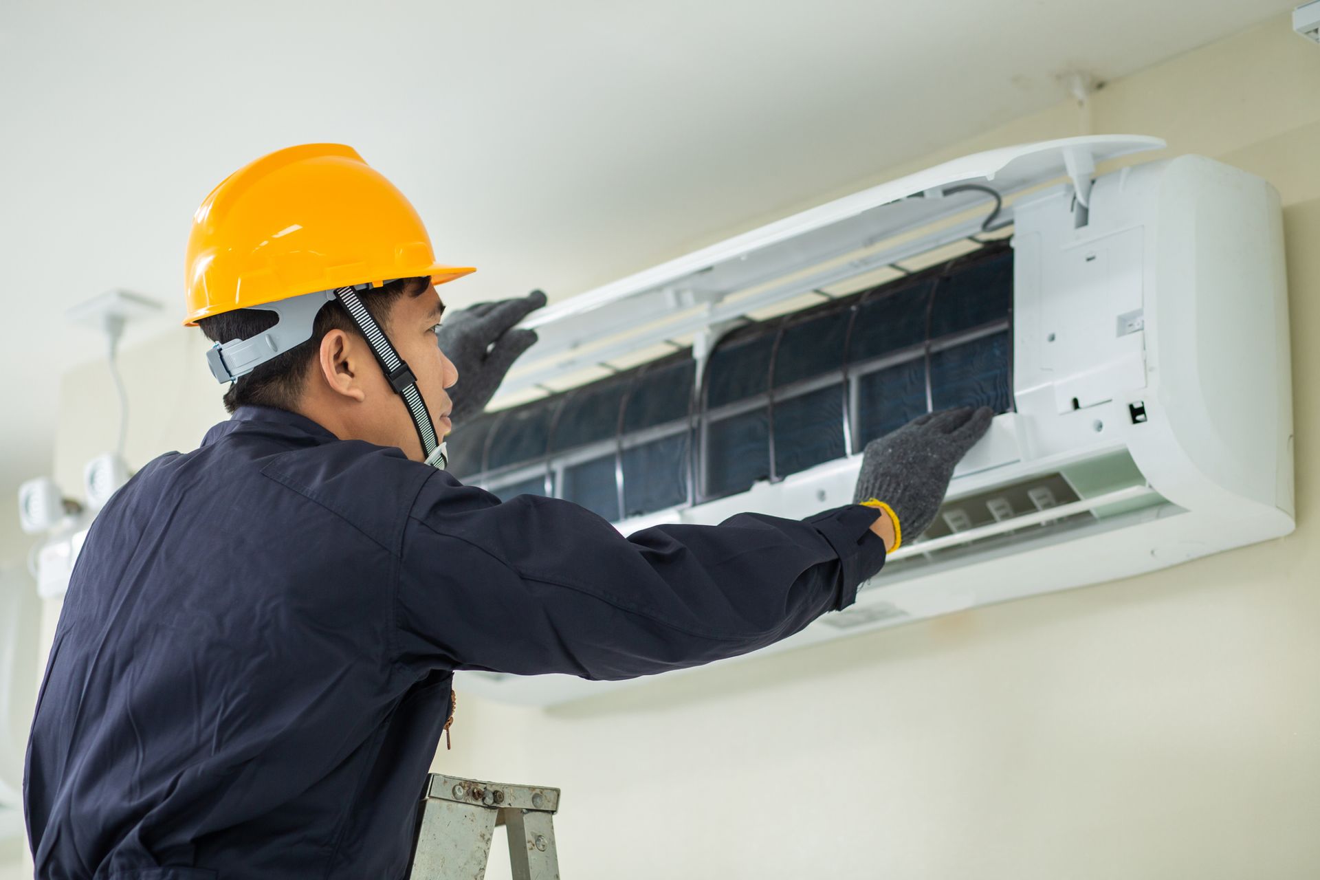 HVAC technician in yellow hardhat inspecting an indoor air conditioning unit, on a wall.