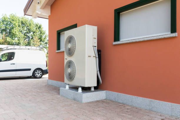 Heat pump unit mounted on a concrete base next to a red building with a service van in the background.