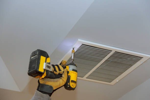 Person using a yellow drill to install a white air vent cover on a white ceiling.