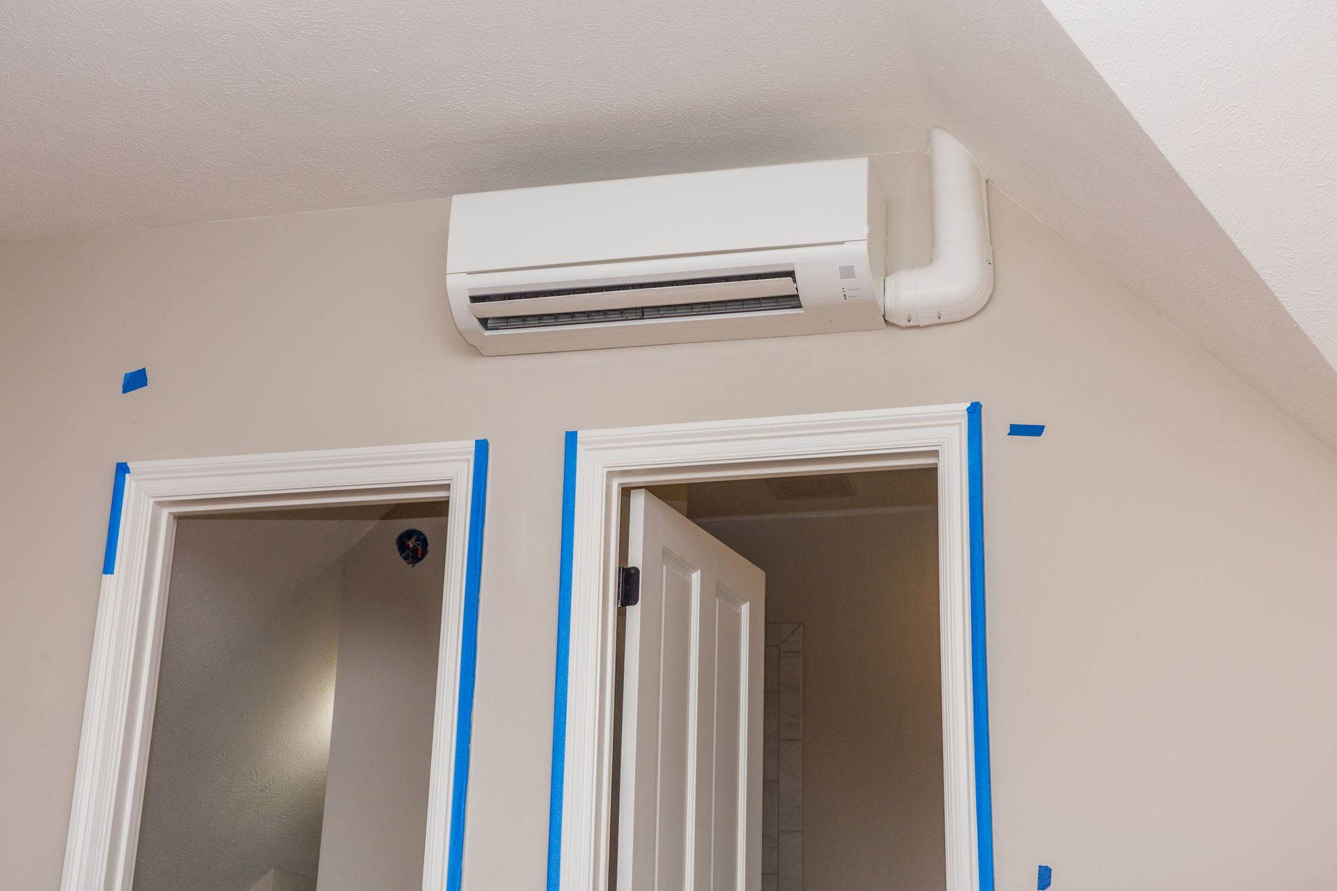 White ductless air conditioner mounted on a beige wall above two white door frames taped with blue painter's tape.