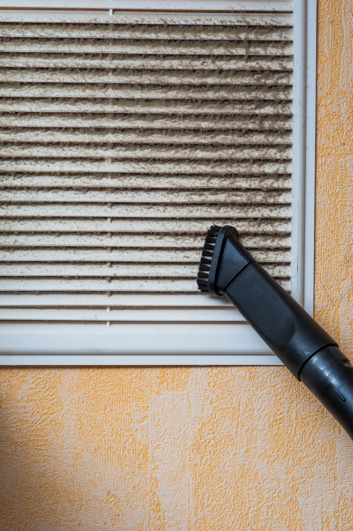 Vacuuming a dirty, dusty air vent on an orange wall.