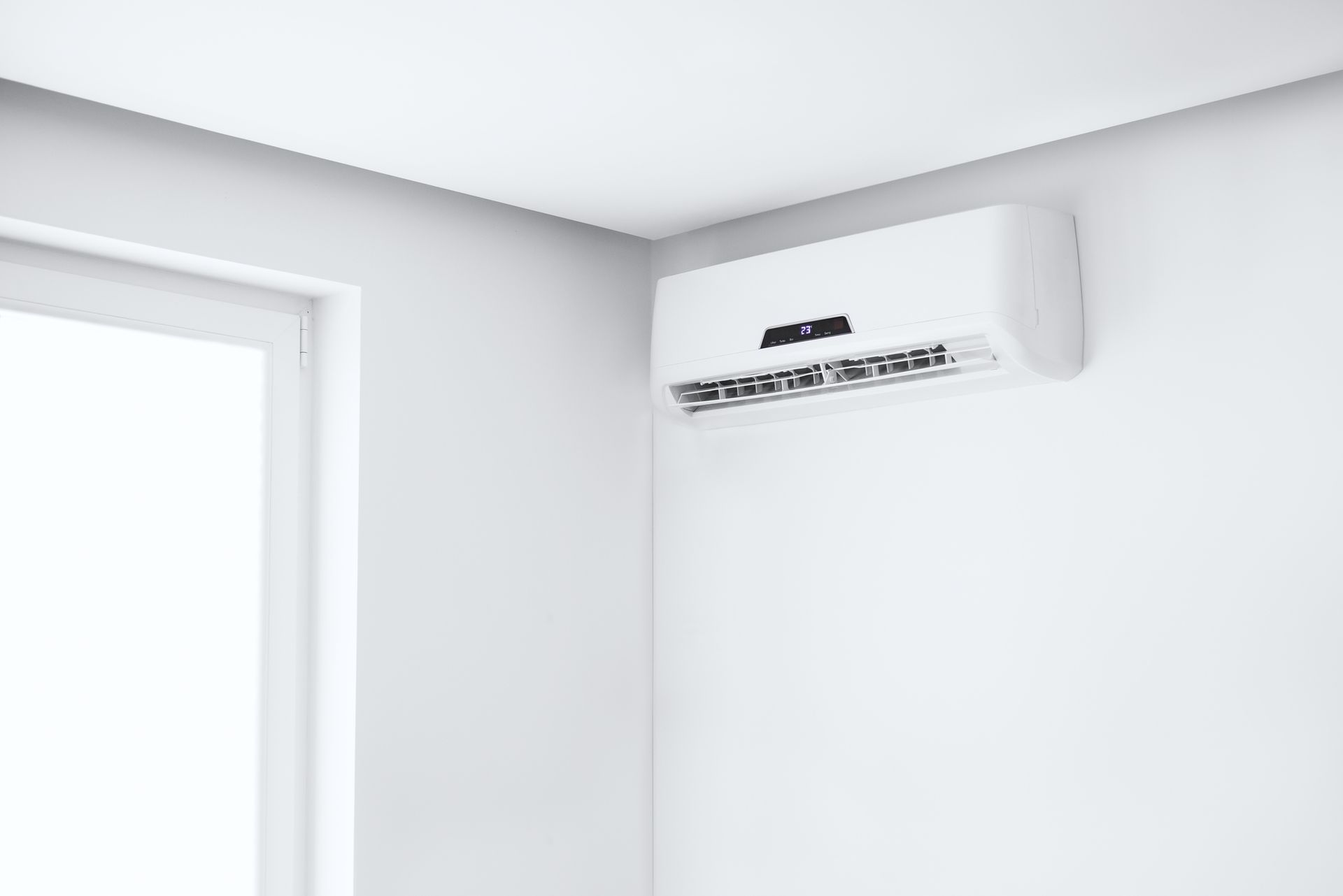 White wall-mounted air conditioner in the corner of a white room with a window.