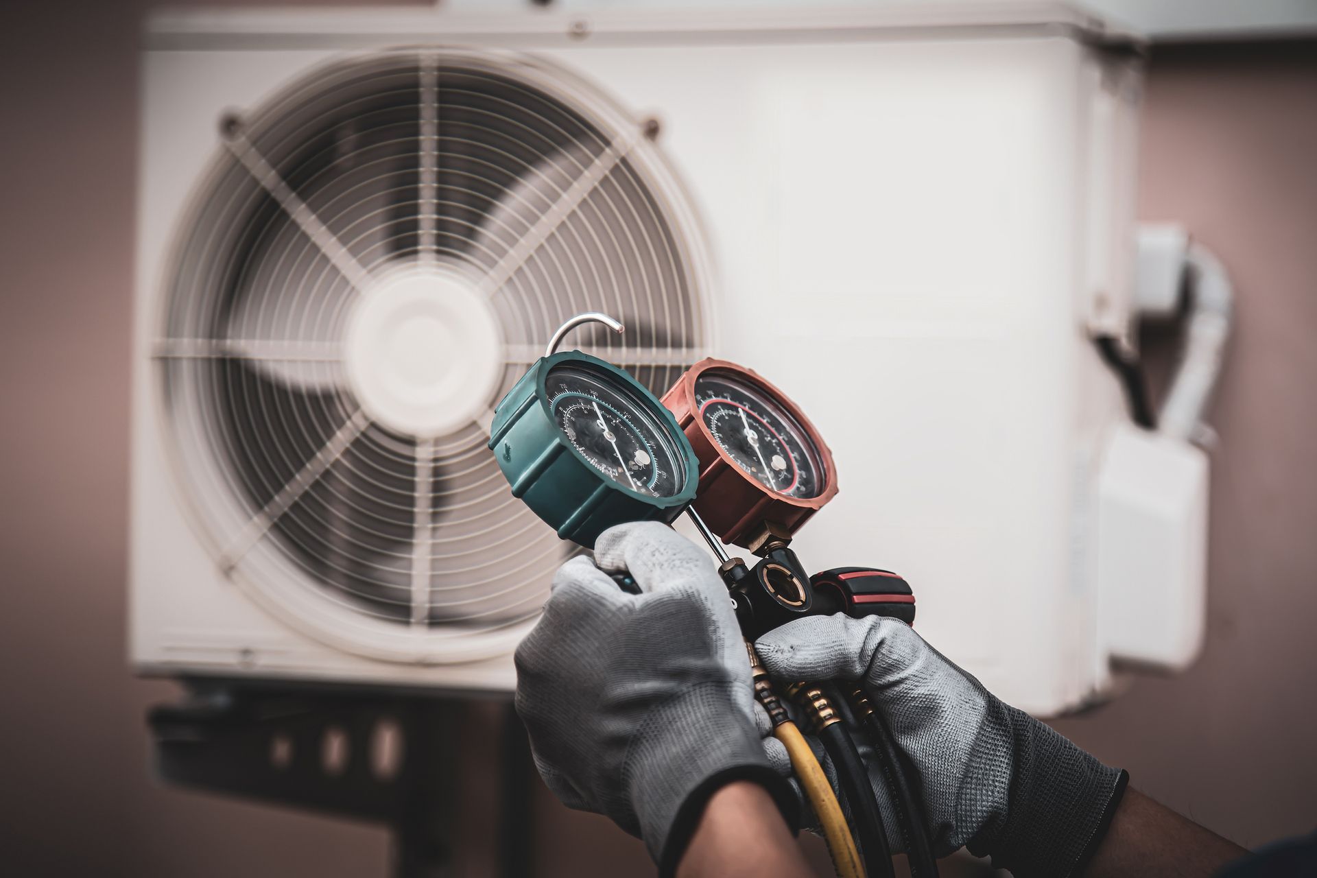 Person wearing gloves holding gauges connected to an outdoor air conditioning unit.