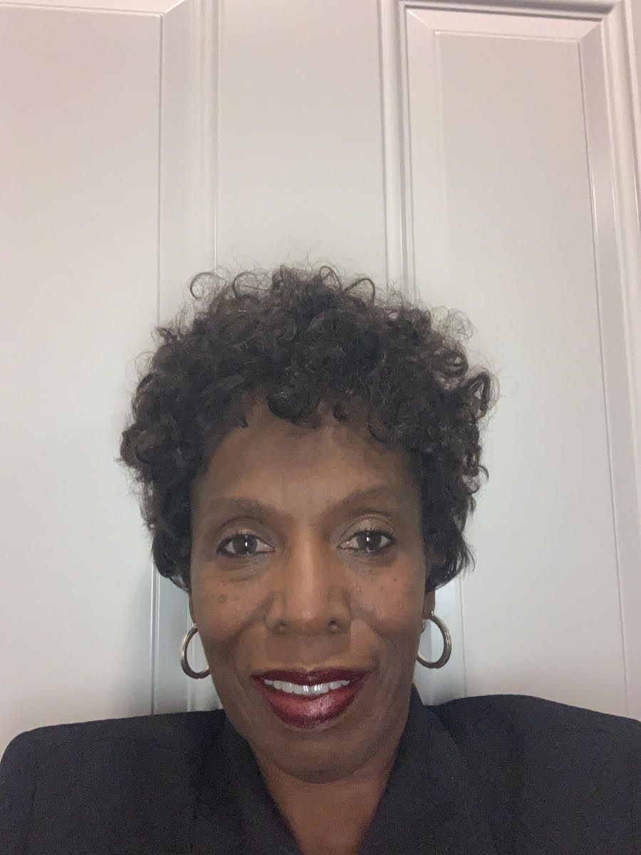 Woman with short curly hair, smiling, wearing a dark blazer, and hoop earrings in front of a white door.