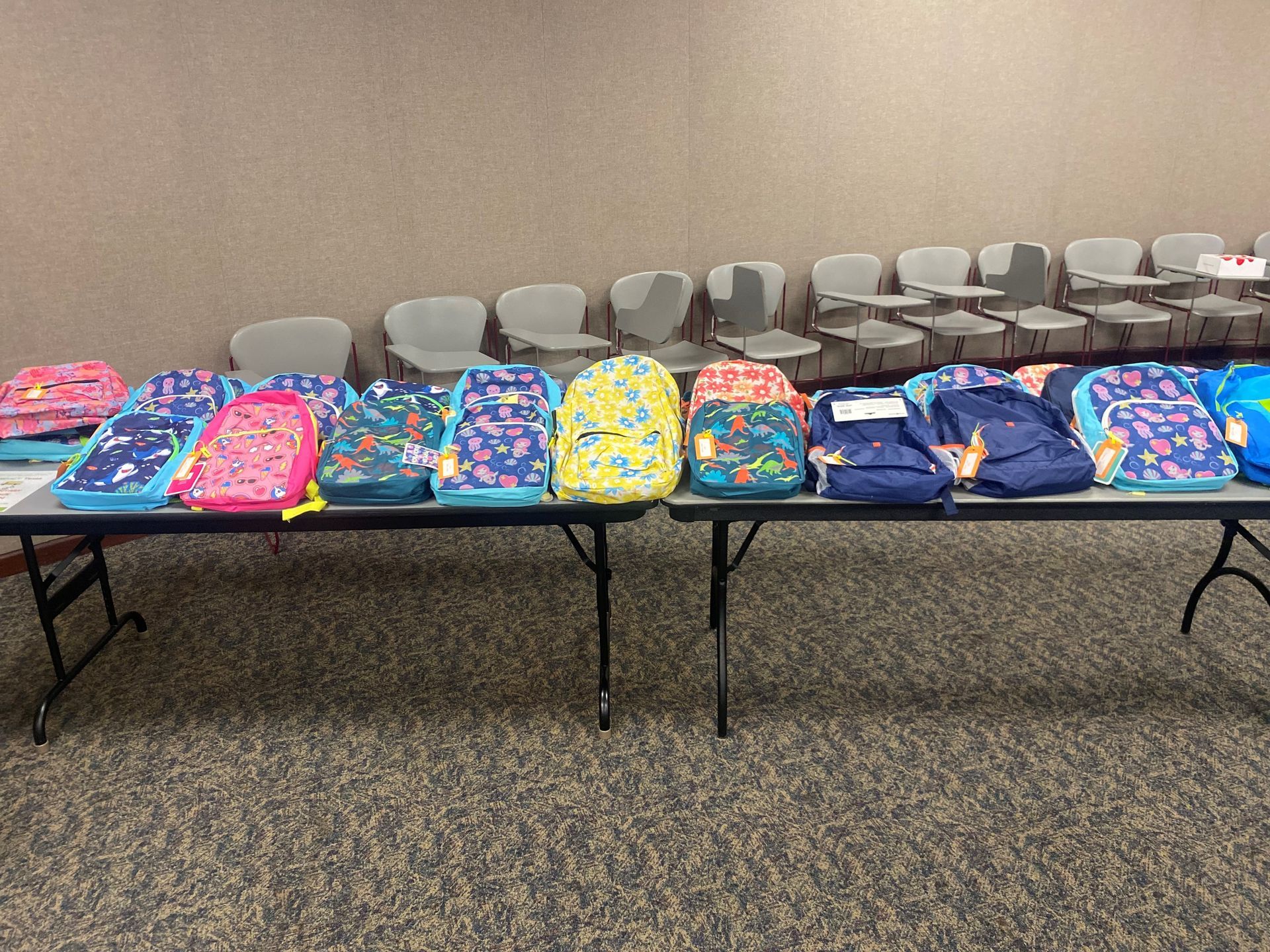 Backpacks on tables in a room with rows of chairs.
