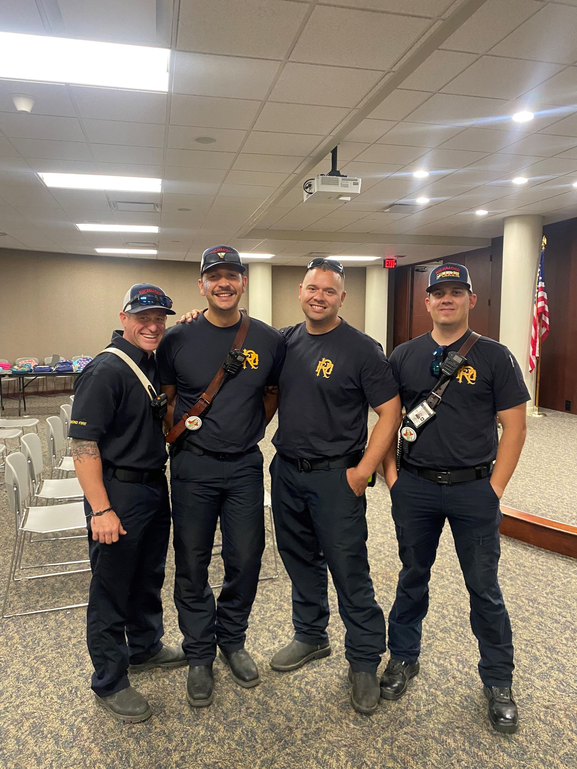 Four people in dark uniforms pose for a photo inside a room with chairs and an American flag.