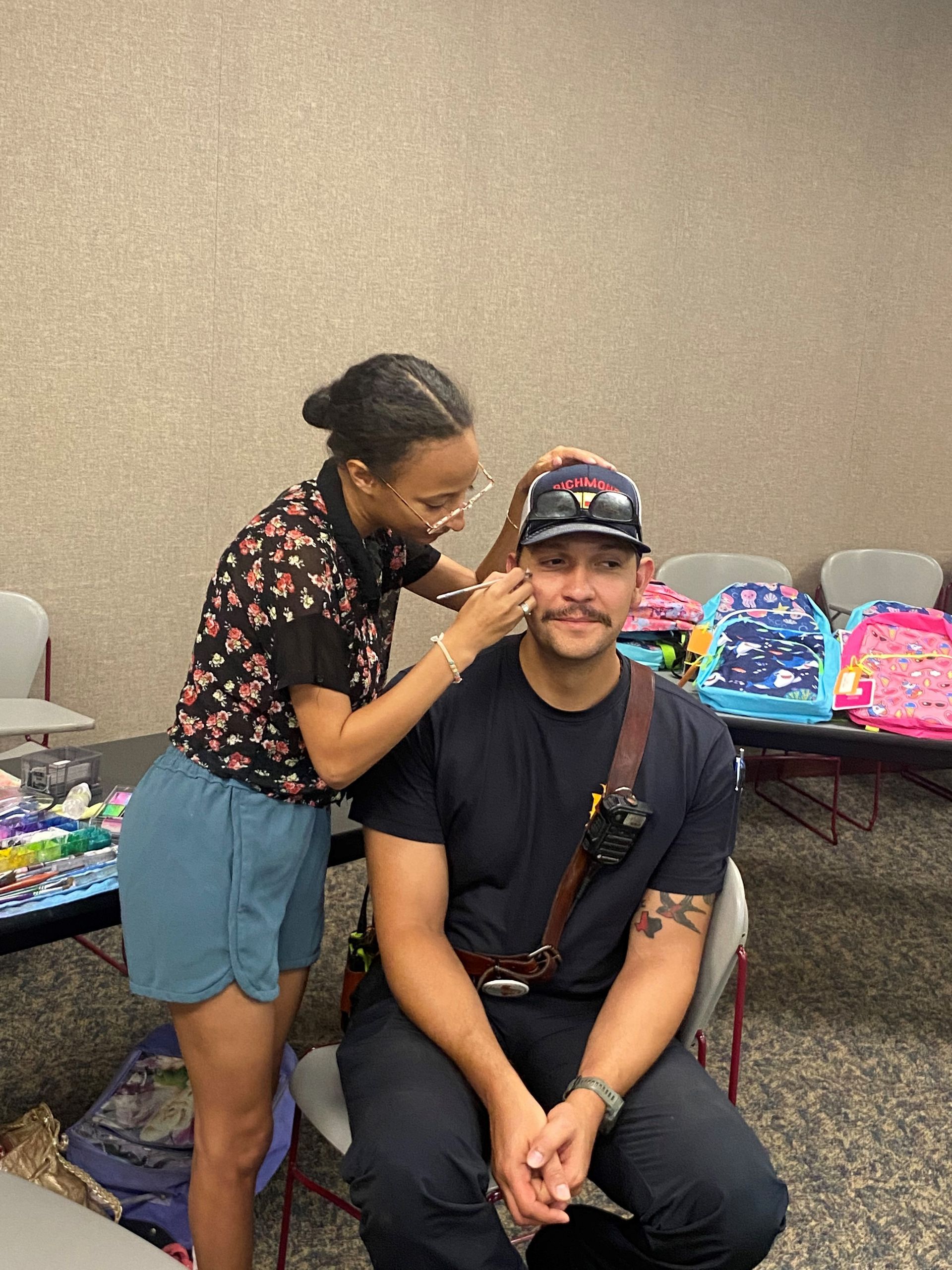 Woman adjusts a hat on a seated man. He's wearing a dark shirt and has a mustache. A table with backpacks is in the background.