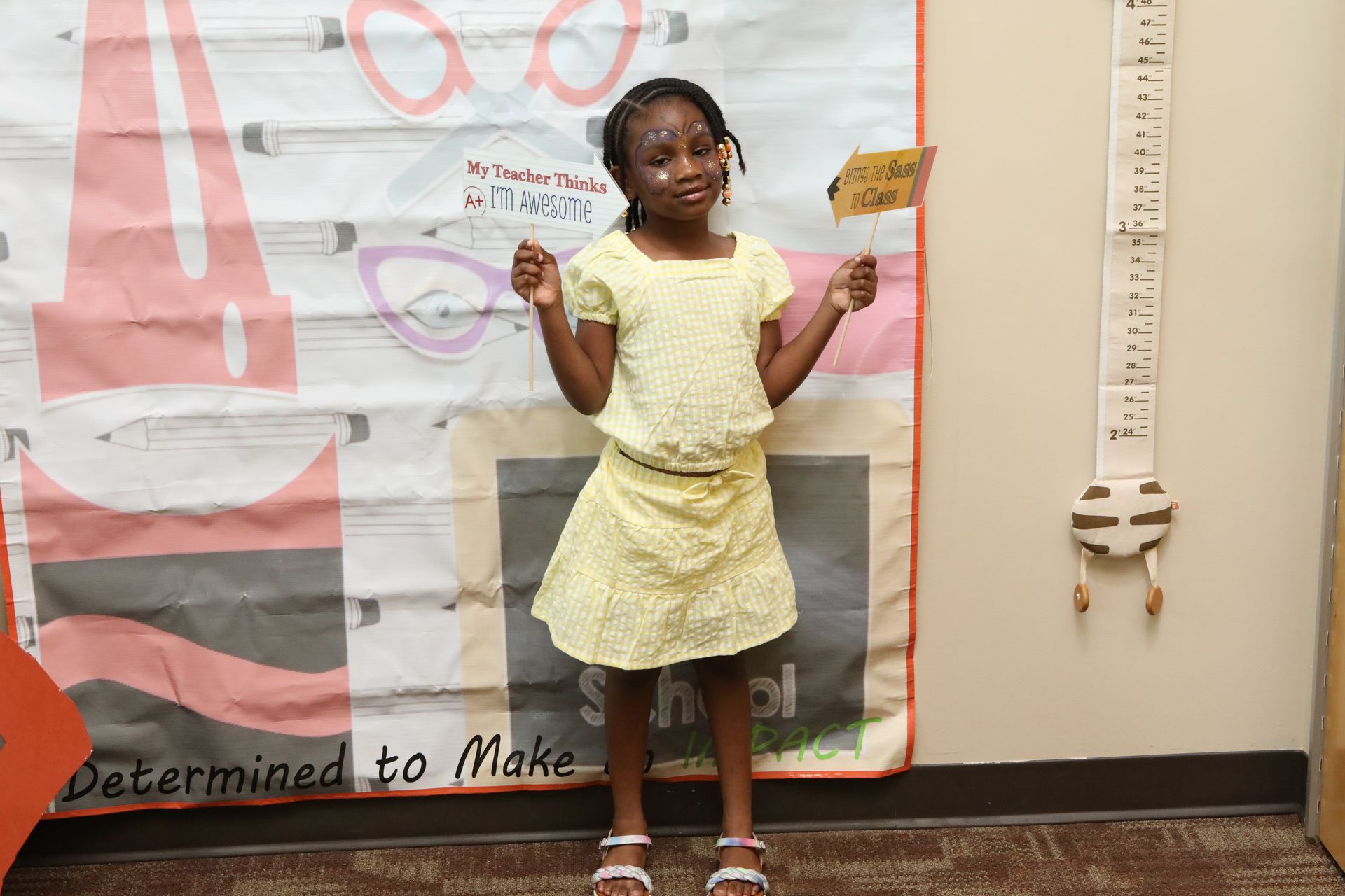 Young girl in a yellow dress holds a sign, standing by a height chart on a wall.