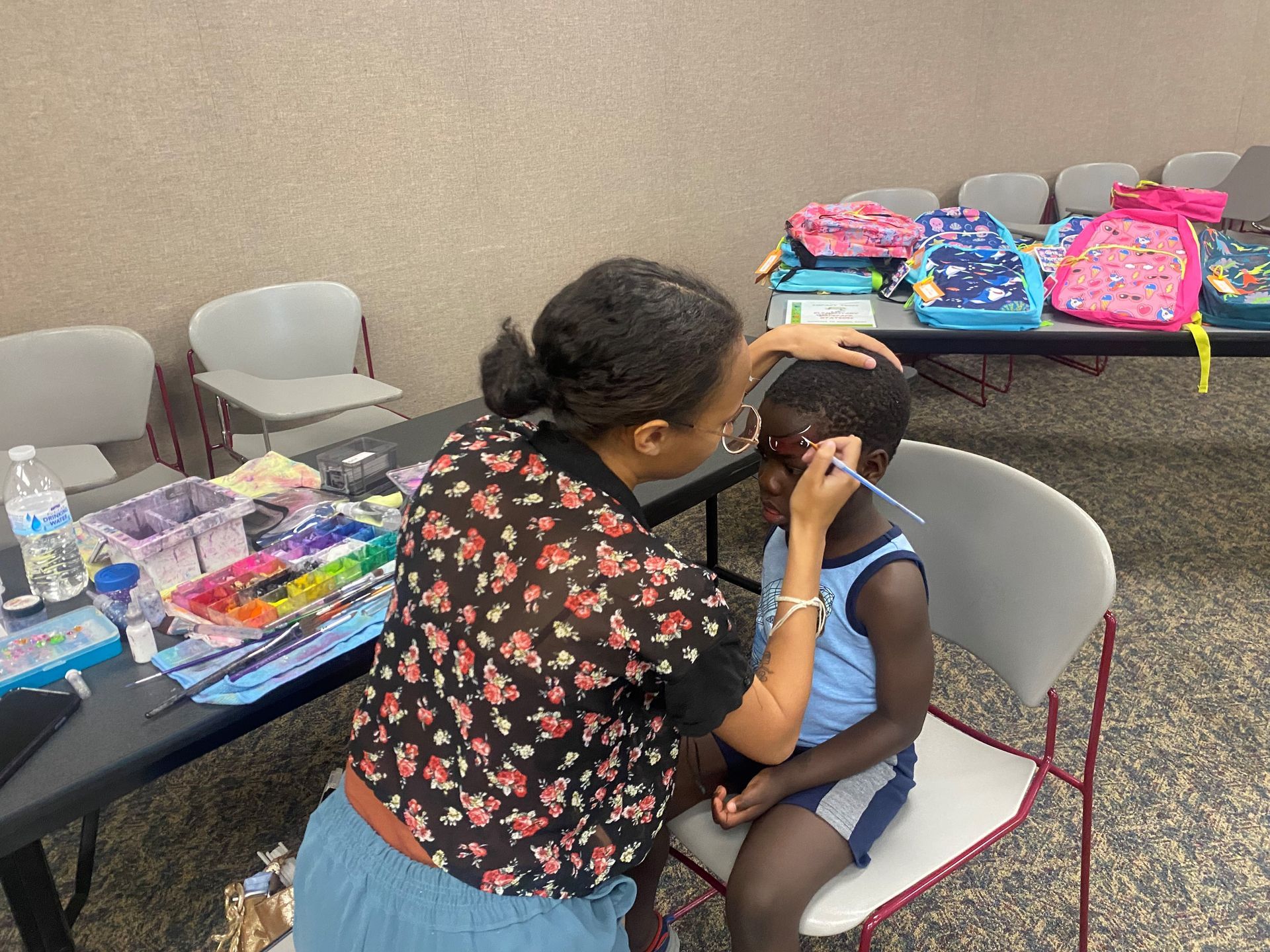 Person painting a child's face at a table with art supplies. Backpacks sit nearby.