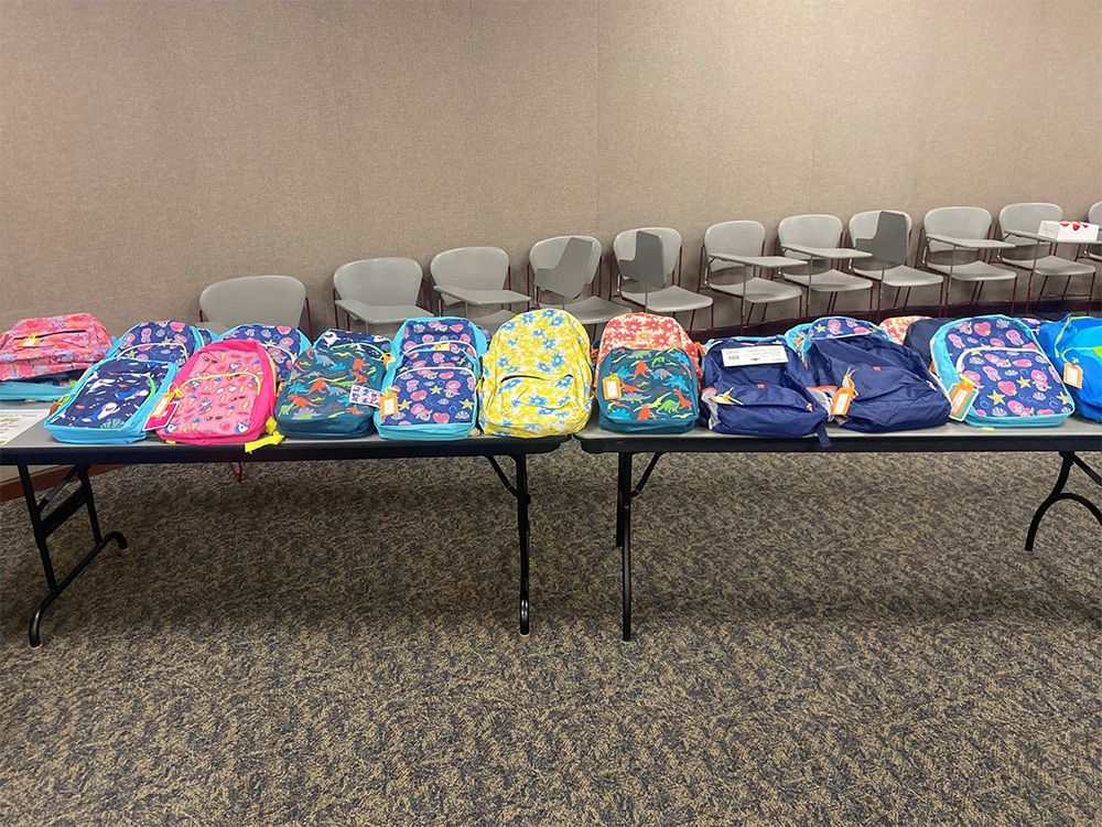 Backpacks of various colors on tables, with chairs in the background.