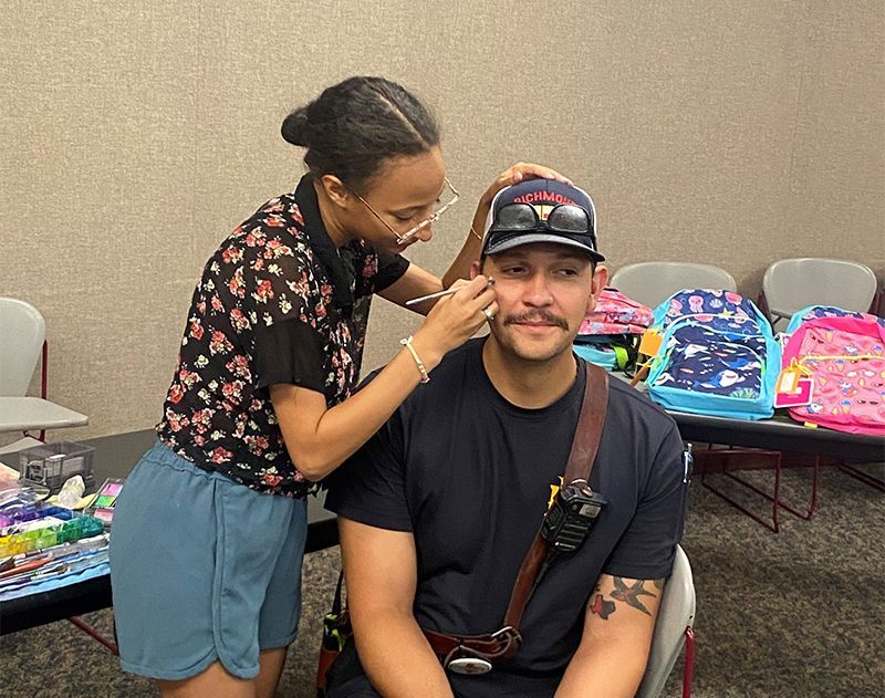 Woman applying face paint to a man's cheek indoors; backpacks in background.