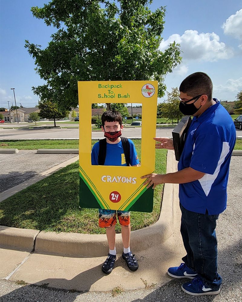 Person posing with a frame cutout. The cutout resembles a crayon box, another person holds it, and a school is in the background.