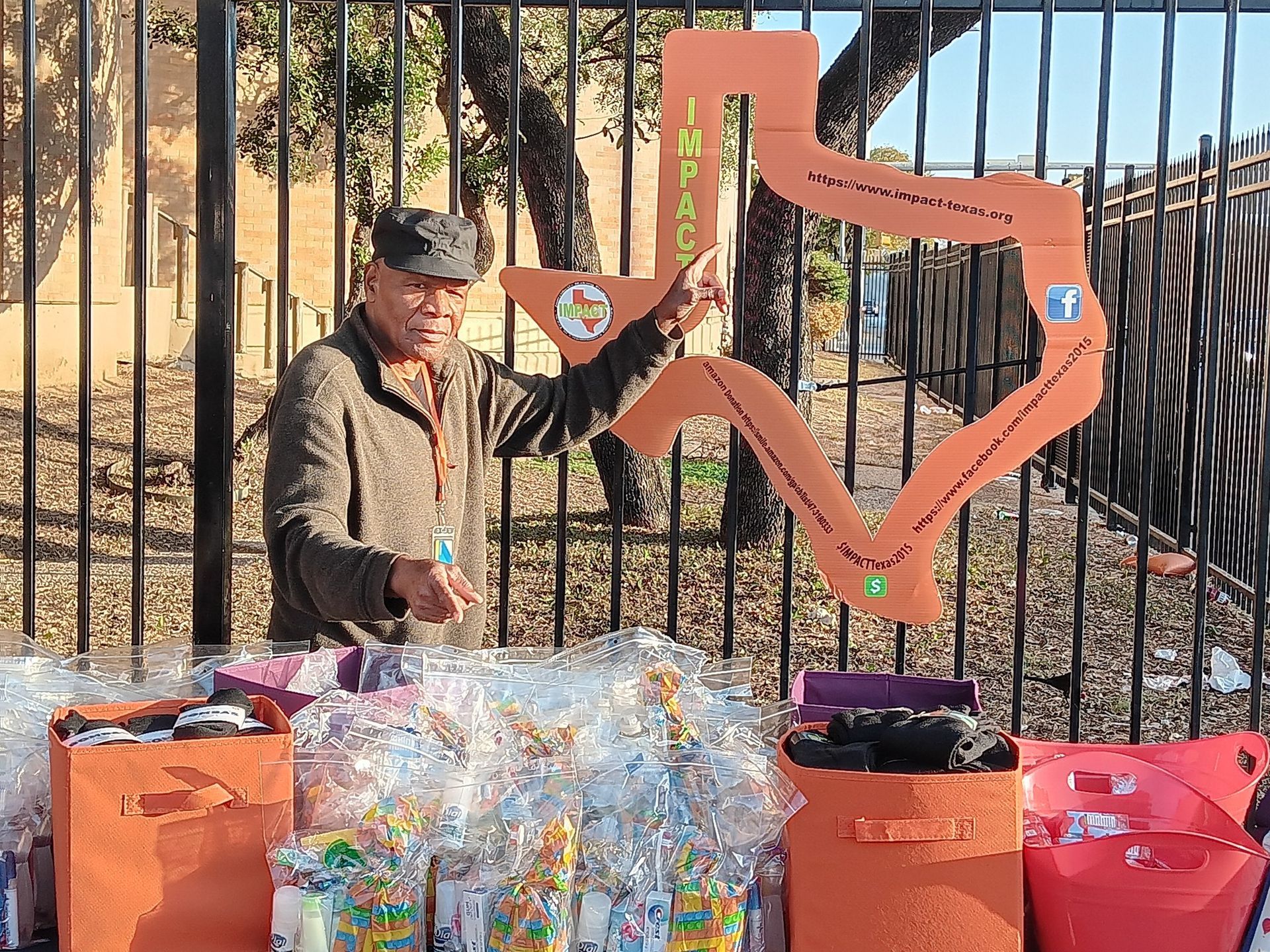 Man points to Texas-shaped sign, selling items from orange bins behind a fence.