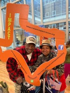 Two people pose with an orange cutout in front of a building. The cutout reads