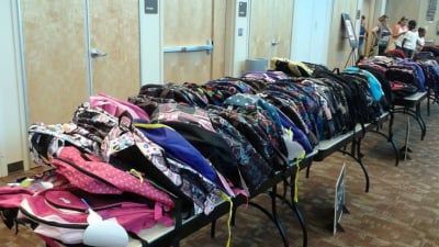 Rows of filled backpacks on tables in a room, ready for distribution.