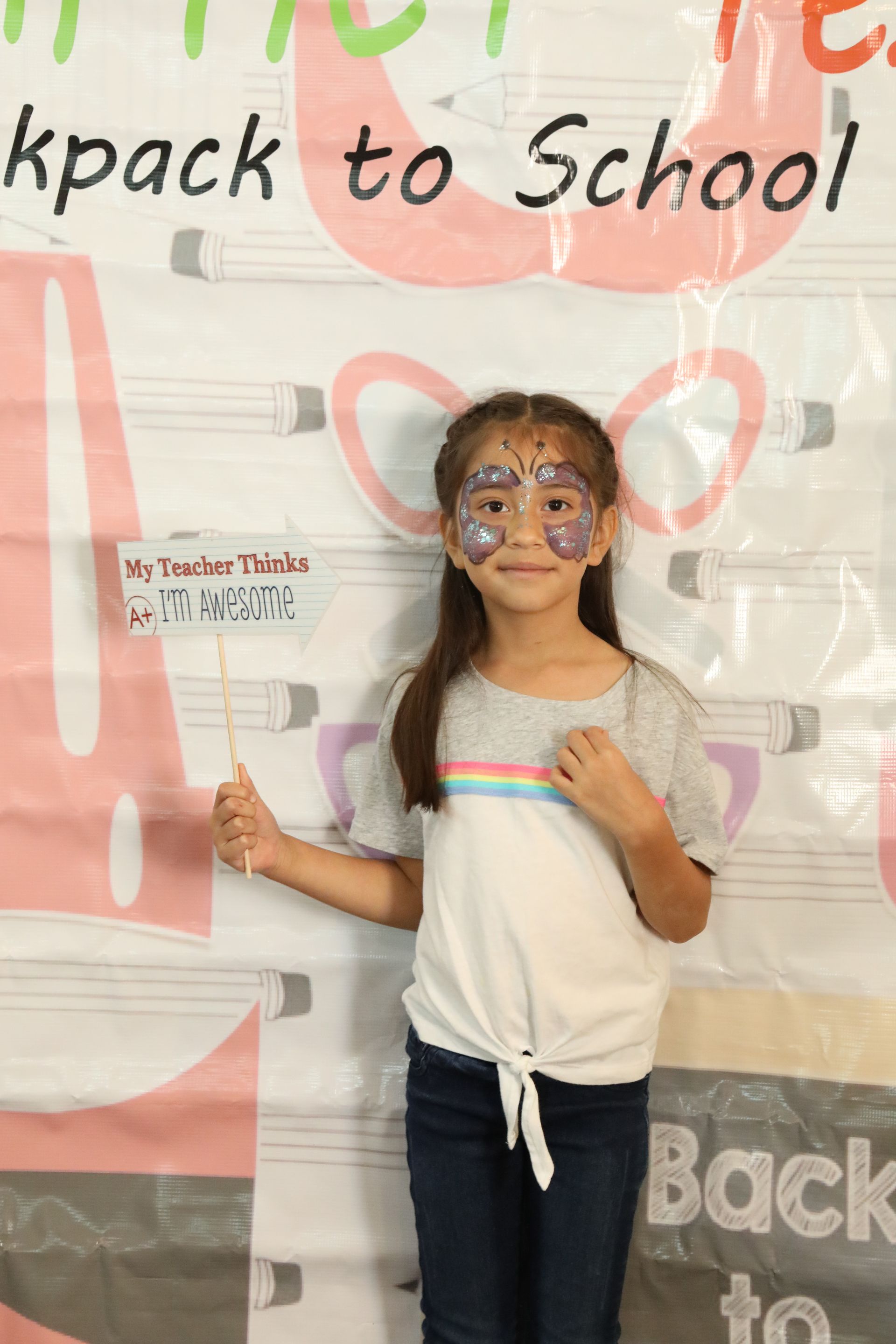 Girl with face paint holding prop, in front of a “Backpack to School” backdrop.