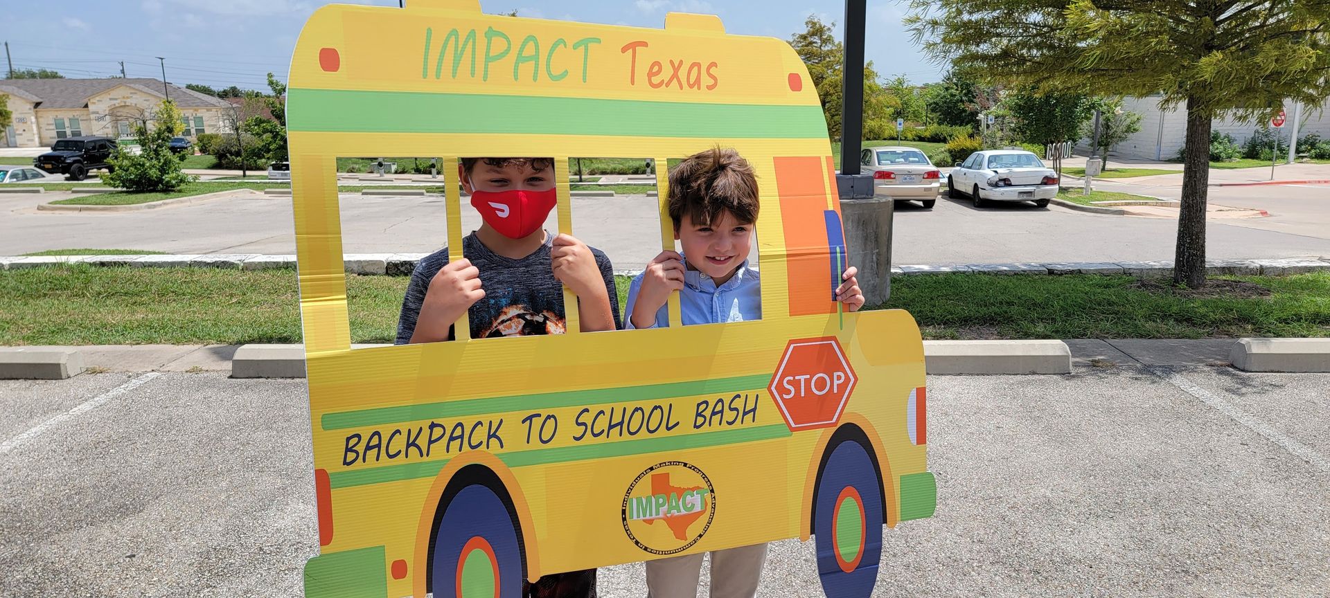 Two children pose in a cardboard school bus cutout. One wears a mask, both give thumbs up, in a parking lot.