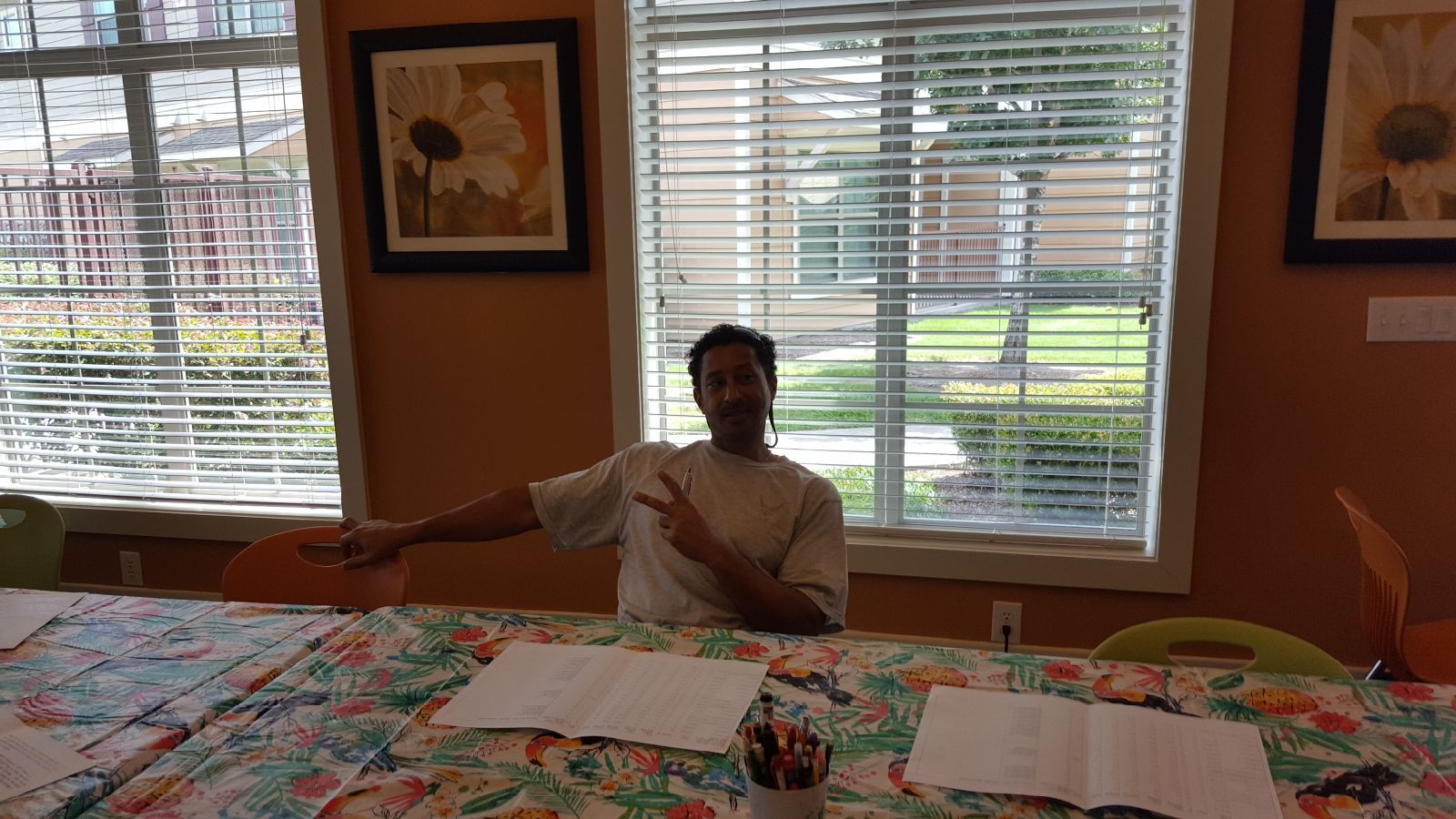 Person at table gestures with arm and hand, papers and pencils, window with blinds in background.