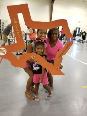 Three people holding a cutout of Texas. Two are smiling and wearing pink. Inside a building, possibly a gymnasium.
