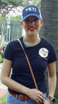 Woman wearing a Yeti hat and a dark T-shirt with a Texas-themed logo. She is smiling outdoors, with a purse.