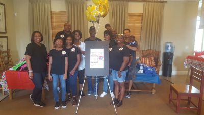 Group of people standing in front of a sign. Yellow balloons are above. Setting appears to be a living room.