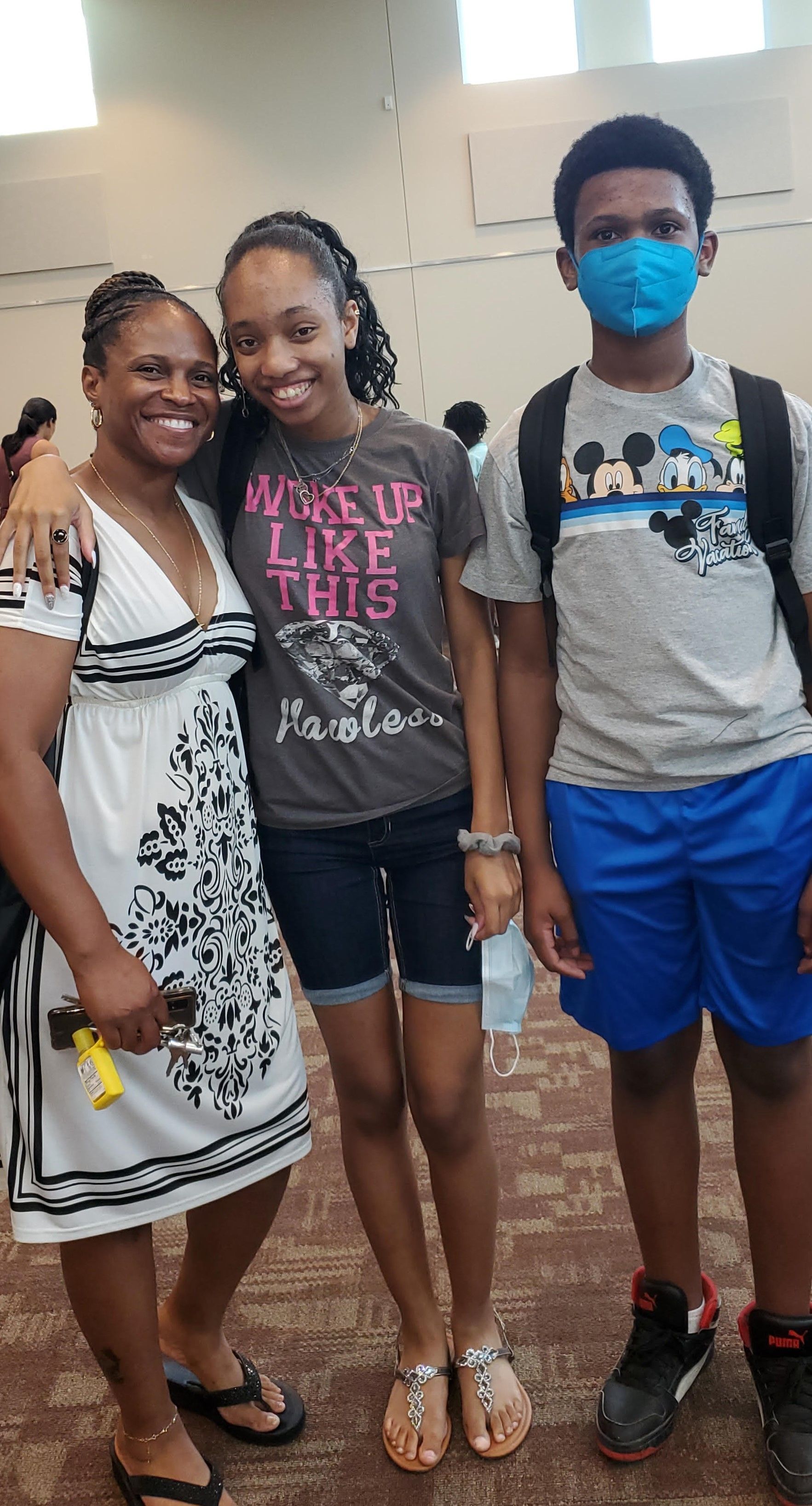 Woman and two teens smiling; woman wearing a white dress, teens wearing casual clothes, indoor setting.