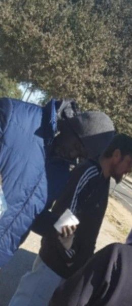 A person in a black jacket and gray hat carries a blue jacket. Holding a white object, standing outdoors.