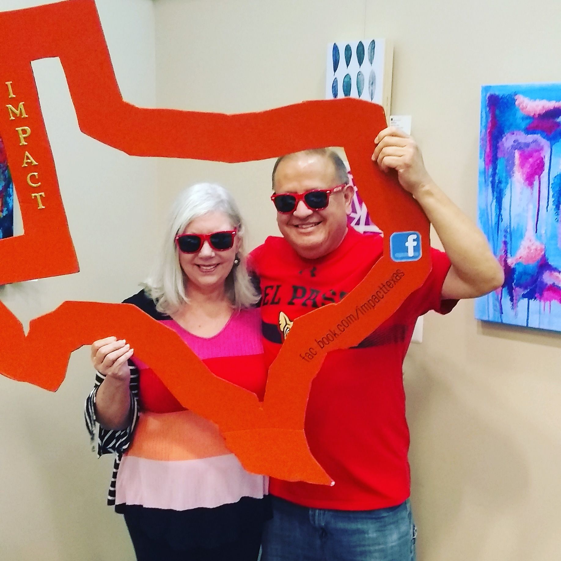 Two people wear red sunglasses, holding an orange cutout that says 