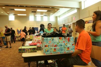 People at a gift distribution event inside a brightly lit room. Boxes filled with items sit on tables.