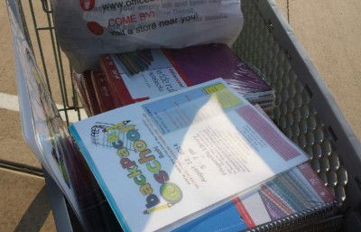 Shopping cart filled with school supplies, including notebooks, folders, and a bag from a store.